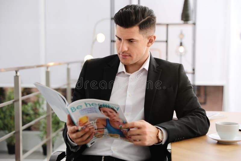 Man in pak leest een gedrukt magazine aan tafel naast een kop koffie